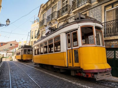 Two vintage yellow and white trams on tracks running along a cobblestone street lined with historic tiled buildings.