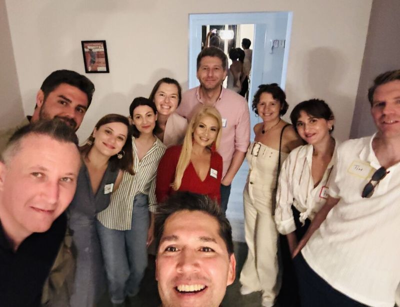 Eric Melchor taking a selfie with a large group of about ten guests wearing name tags at a Bucharest cocktail party