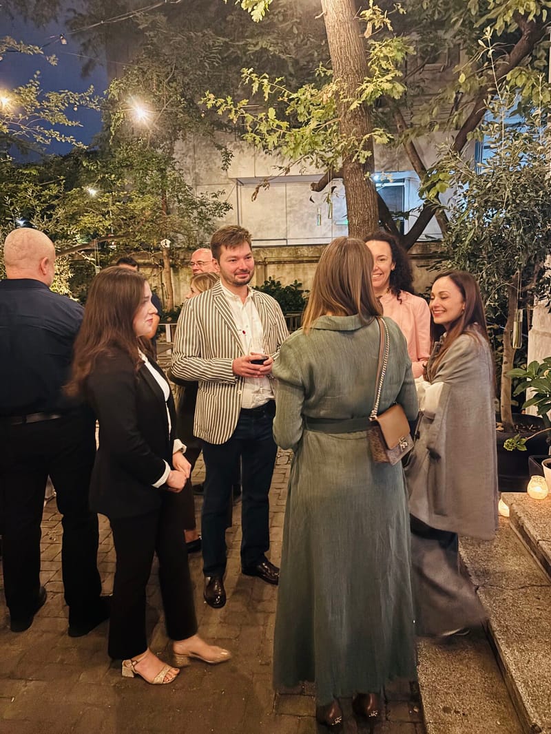 Group of guests chatting under string lights in a tree-lined courtyard at dusk, a man in a striped blazer at the center of the conversation