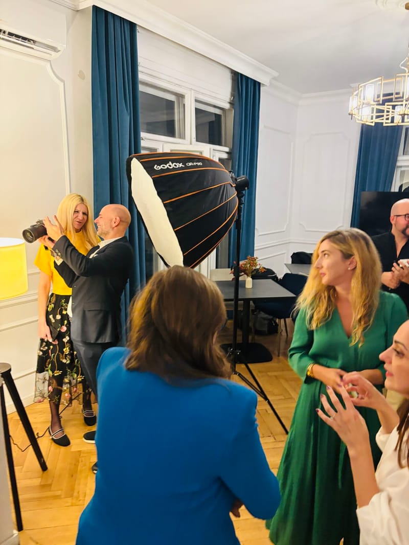 A professional photographer with a large softbox light photographs a woman in a yellow top and floral skirt while other guests watch in an elegant room