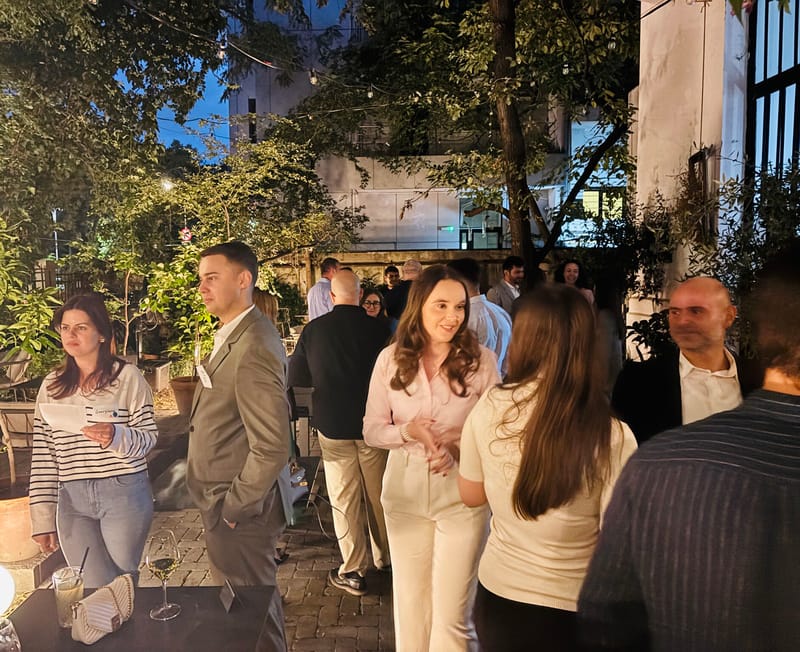 Outdoor evening scene with guests mingling on a patio, string lights overhead, a woman in pink at the center talking with others