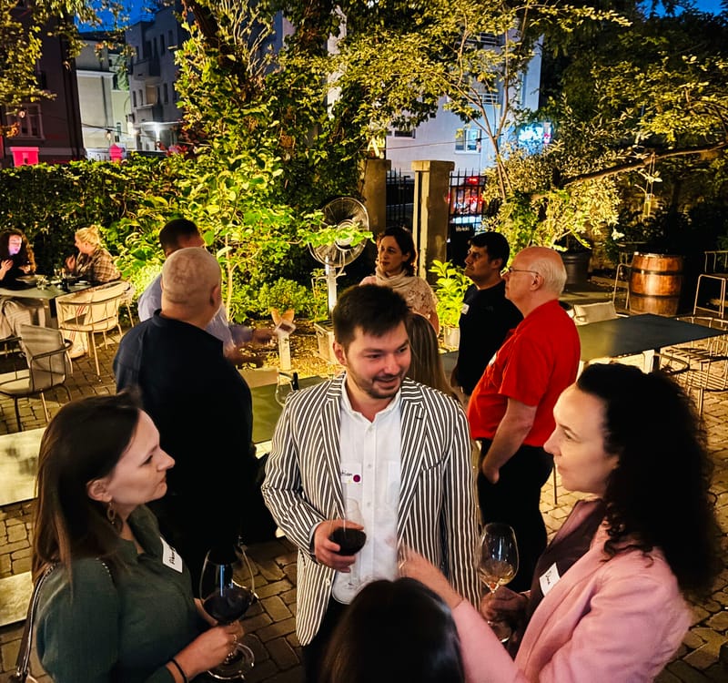Evening garden party with guests mingling under string lights, holding wine glasses, surrounded by trees and warm ambient lighting