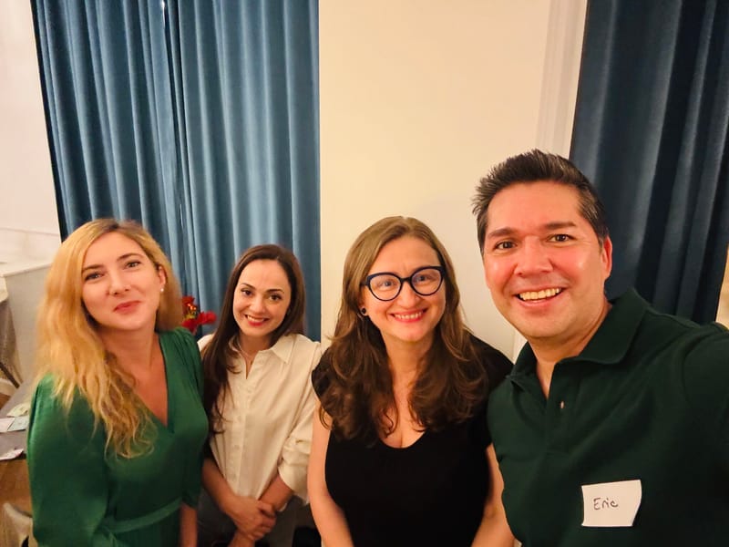 Four people smiling in a selfie at a party, three women and one man wearing a green polo with a name tag, blue curtains behind them